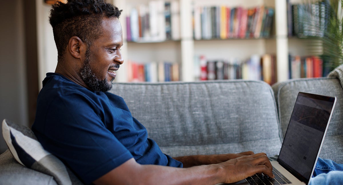 Smiling black man sits on sofa working on open laptop
