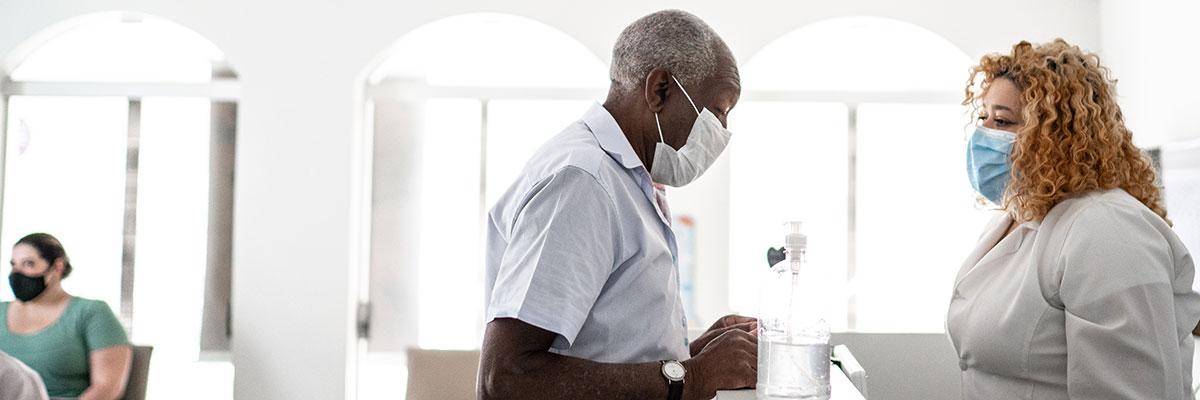 Black elderly patient in medical mask stands at medical registration desk speaking with younger attendant, also masked